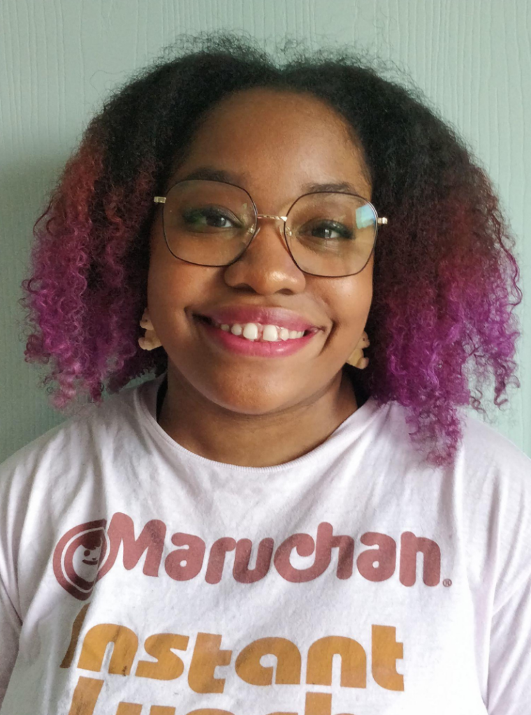 A brown-skinned Black woman with curly hair dyed a vivid magenta at the ends. She wears glasses and a bright smile, along with a pretty cool Instant Ramen shirt.